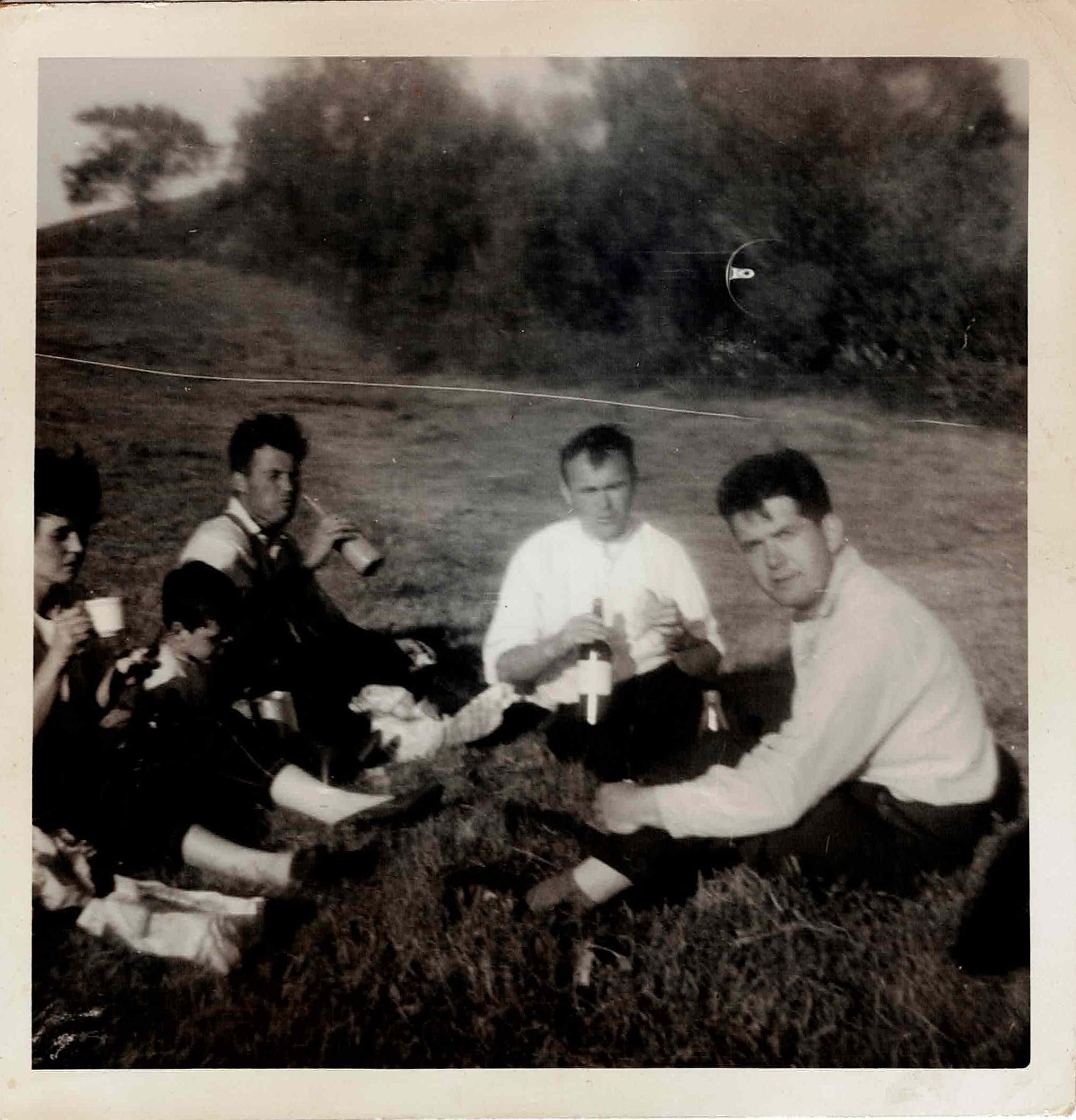 a black and white photo taken in
    the 1960s showing three men in their 20s-30s, a young boy, and a woman in
    her late 20s. the men are drinking from glass bottles. the young boy is
    drinking from a tin mug, a 'pandy'.